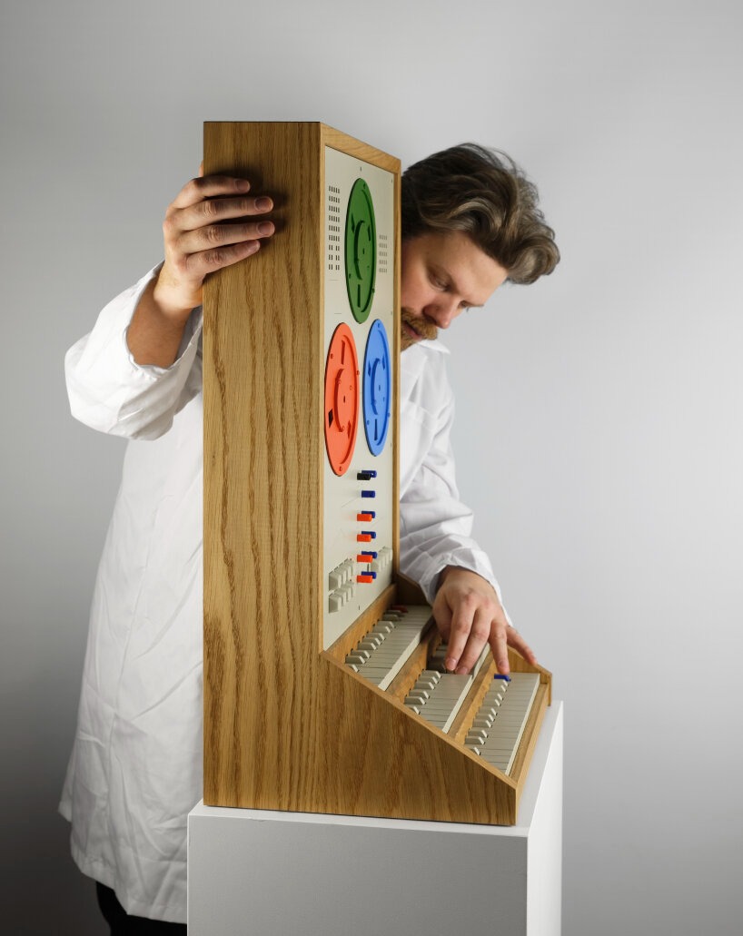 A person in a white lab coat interacts with a large, wooden, vintage-style electronic device featuring colorful dials and buttons.