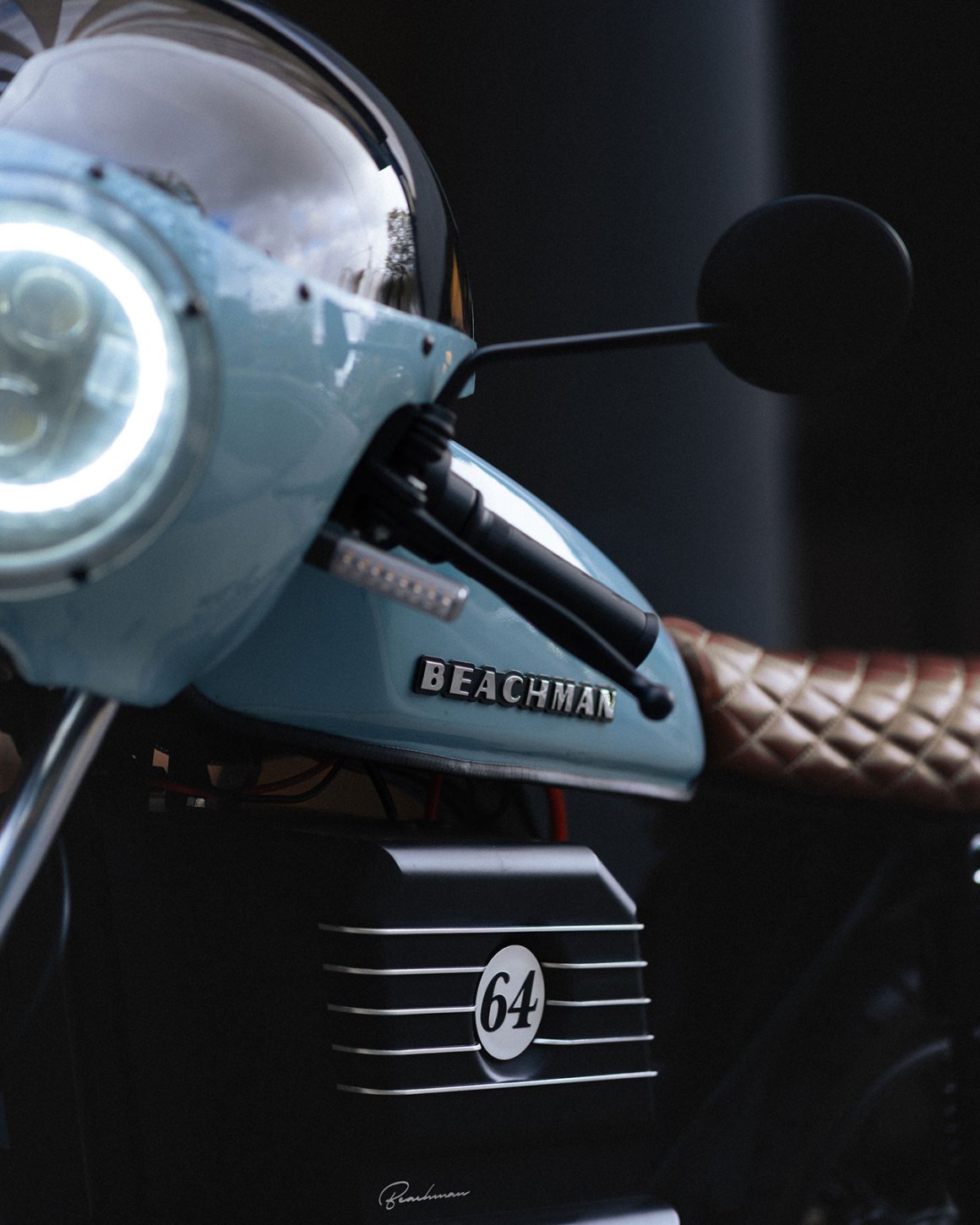 Close-up of a vintage motorcycle with a light blue body, featuring the name 'BEACHMAN' on the front, a round headlight, and a distinctive tan quilted seat design.