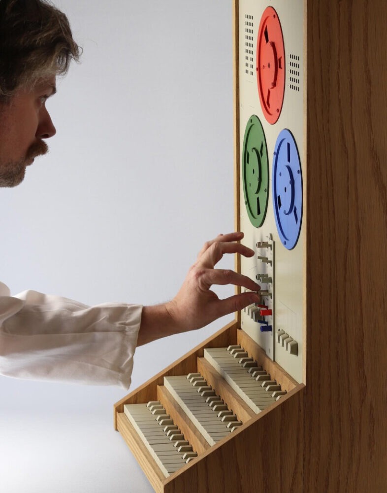A person in a white shirt interacting with a colorful control panel featuring four circular dials and a series of buttons, with a wooden structure beneath that includes rows of keys.
