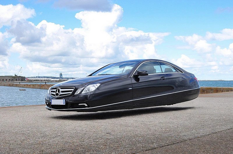 A digitally edited image of a black Mercedes-Benz car with a smooth, hovering undercarriage, set against a blue sky with fluffy clouds and a body of water in the background.