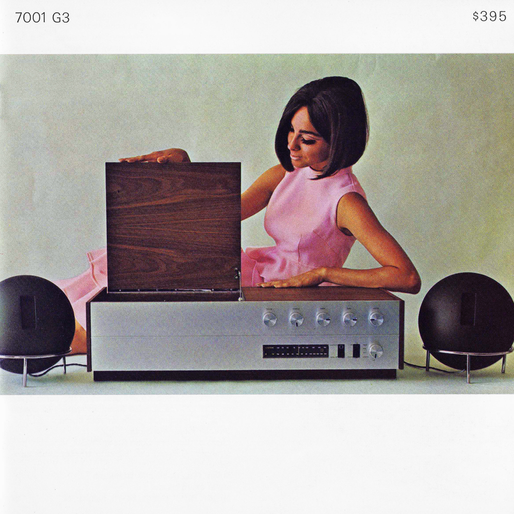 A woman in a pink dress sits beside a vintage audio system with a wooden lid, featuring knobs and dials, flanked by two round speakers.