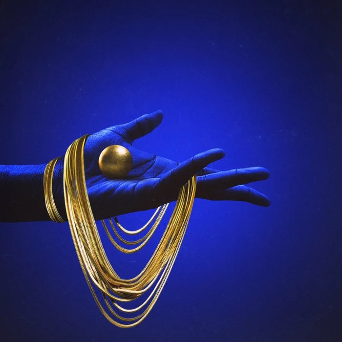 A blue hand holding a golden sphere with multiple strands of gold jewelry draped over it against a blue background.