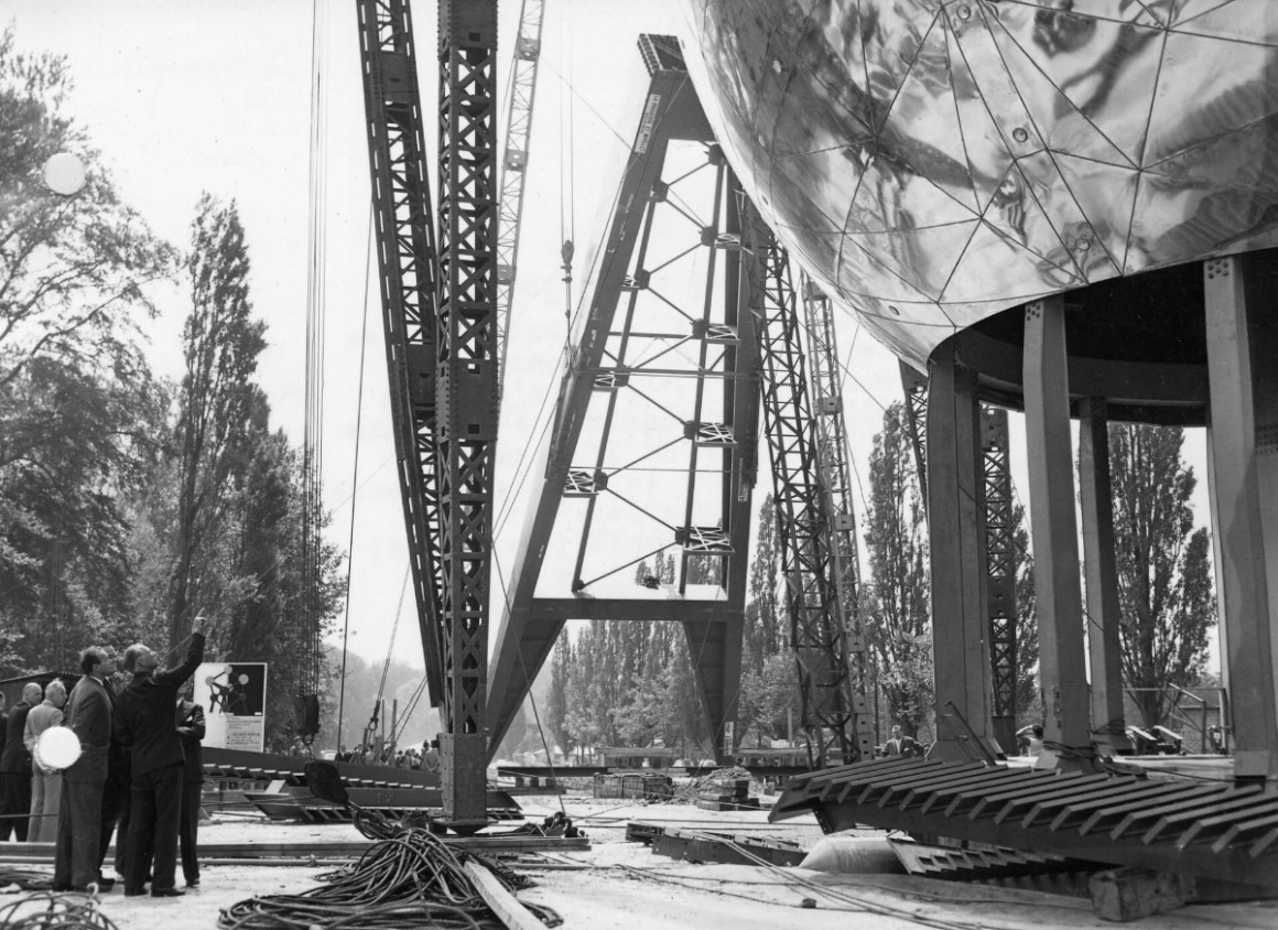 Black and white photo of the construction of the Atomium in Brussels, 1958. Several workers observe large steel beams and supports being assembled, with cranes and trees in the background.
