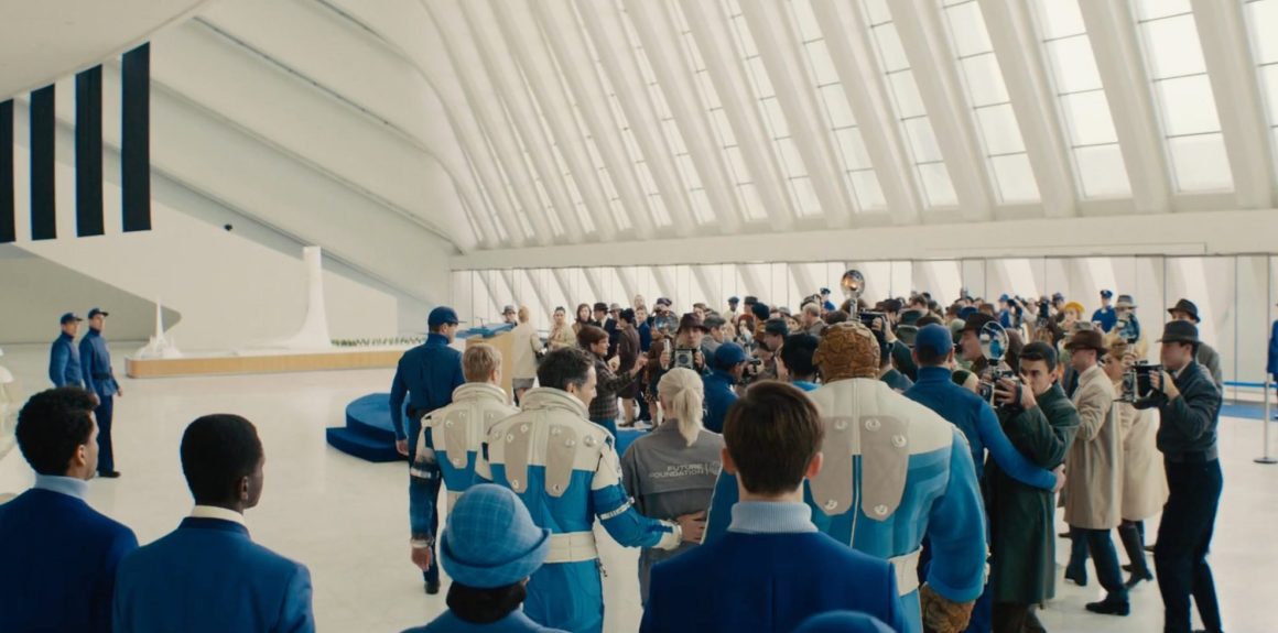 A crowded indoor space with people dressed in blue outfits, some wearing caps, and others in formal attire, gathered around a central figure. The architecture features large, curved white structures and natural light coming through expansive windows.