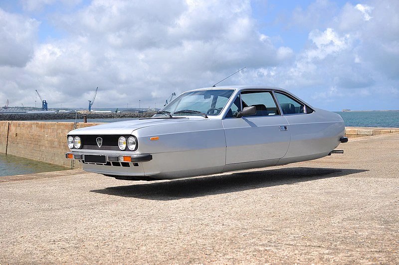 A silver car appearing to hover near a dock, with a cloudy sky and water in the background.