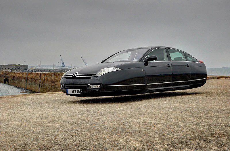 A black Citroën vehicle appears to be floating above a concrete surface near a harbor under a cloudy sky.