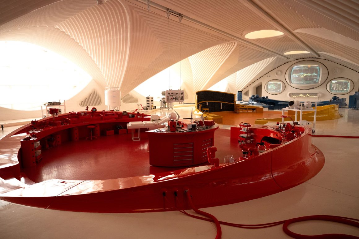 An interior view of a modern laboratory space featuring a curved red workstation, surrounded by unique architectural elements and various instruments in a bright environment.