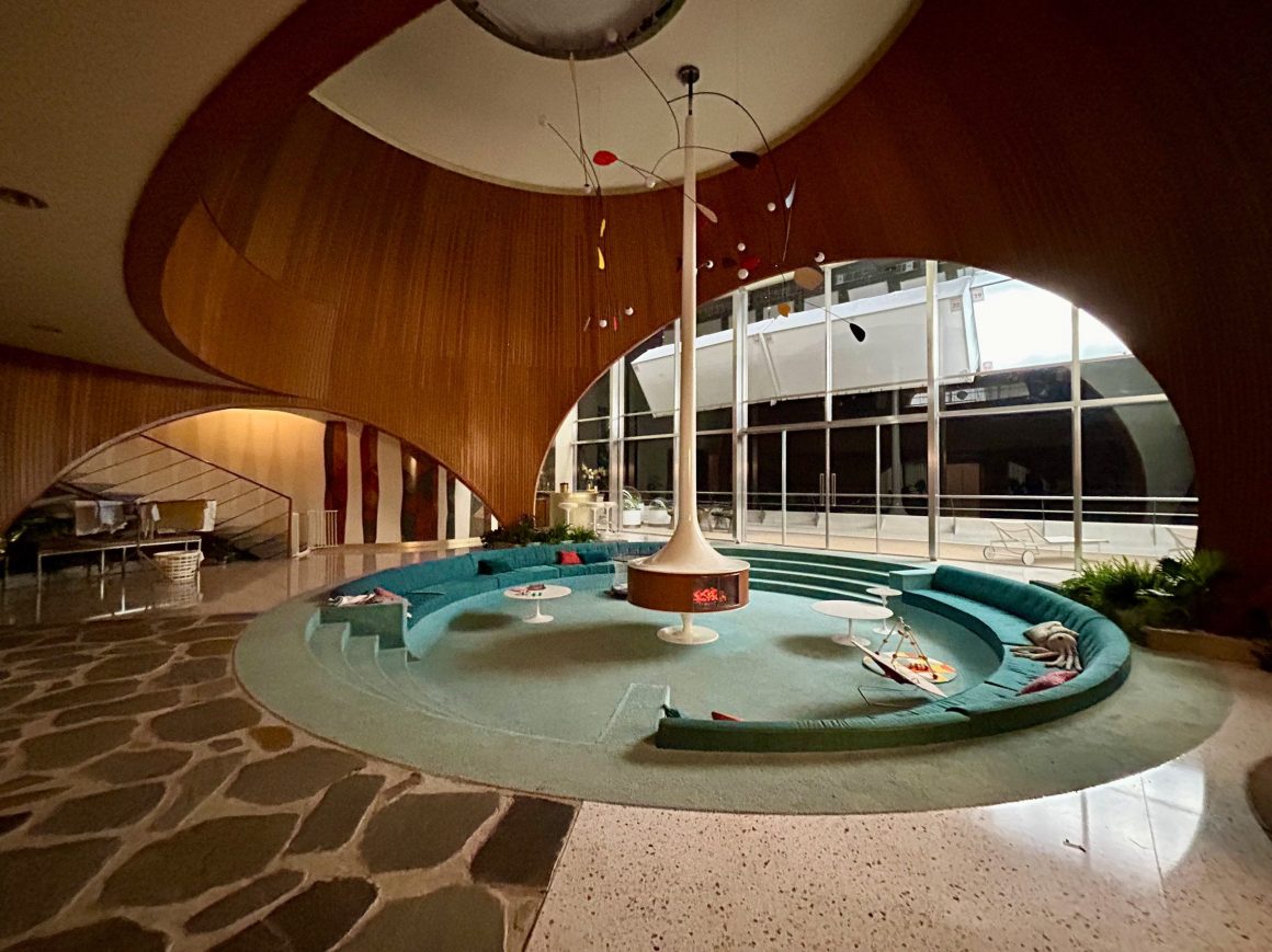 Interior of a modern, curved lounge area with circular seating, featuring a central column and large windows, showcasing natural light.