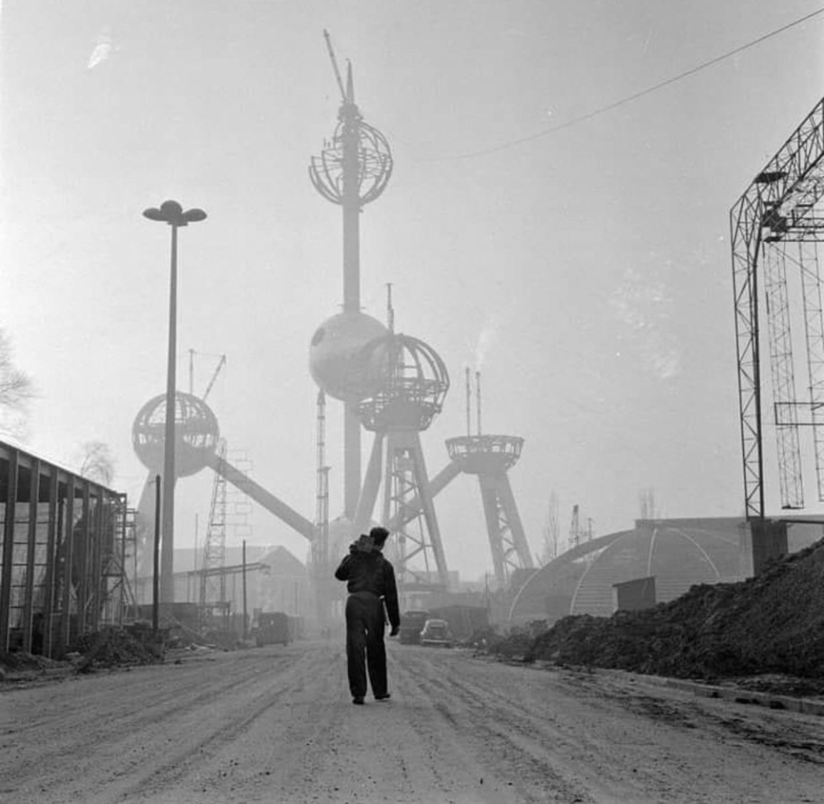 A black and white photo of a man walking down a dirt road, with the Atomium structure in the background, partially under construction, surrounded by scaffolding and buildings.