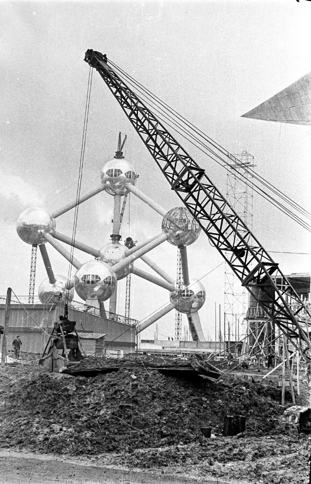 Black and white photo of the Atomium under construction, showcasing a crane in the foreground and the gleaming spheres of the structure behind.