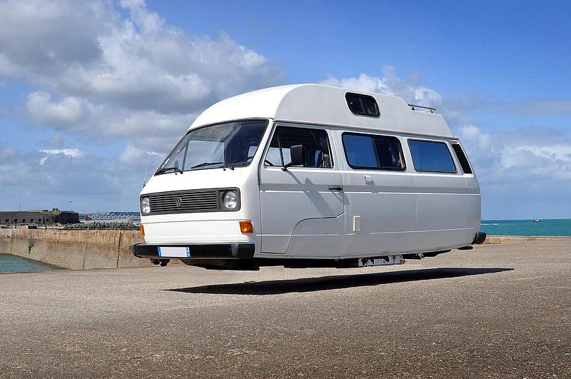 A white camper van hovering above a concrete surface with a blue ocean and cloudy sky in the background.