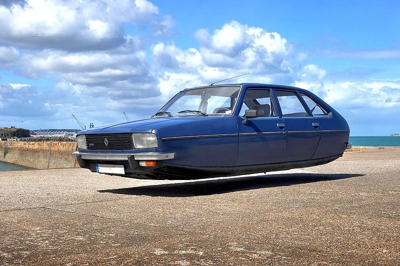 A blue car appears to be levitating above a concrete surface with a beach and cloudy sky in the background.