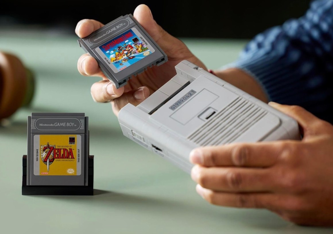 A person holding a Nintendo Game Boy game cartridge with the label 'Super Mario Land' while inserting it into a Game Boy console, with another cartridge for 'The Legend of Zelda: Link's Awakening' displayed on a stand.