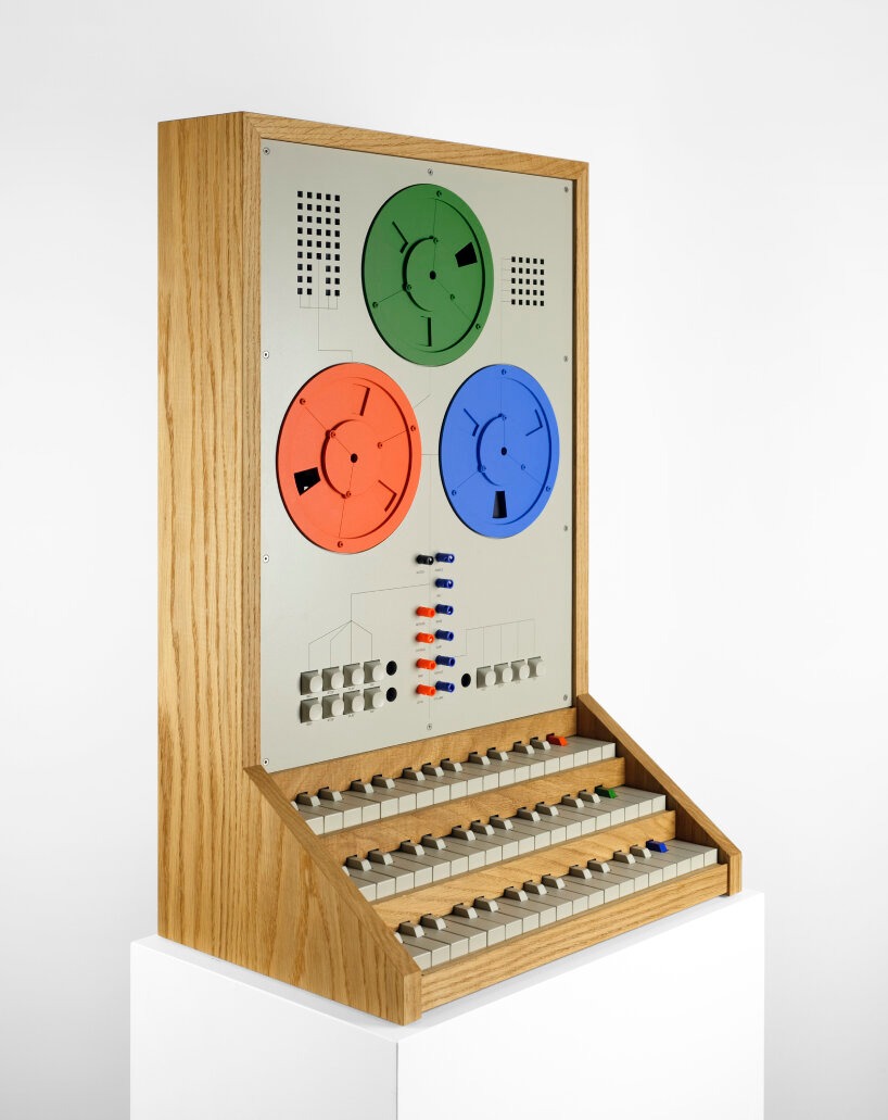 A vintage electronic synthesizer with colorful circular dials and an array of keys, presented on a white pedestal against a plain background.