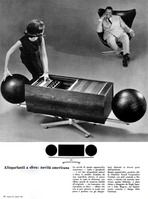 A woman is interacting with a unique audio equipment featuring spherical speakers, while a man sits relaxed in a modern chair in the background.