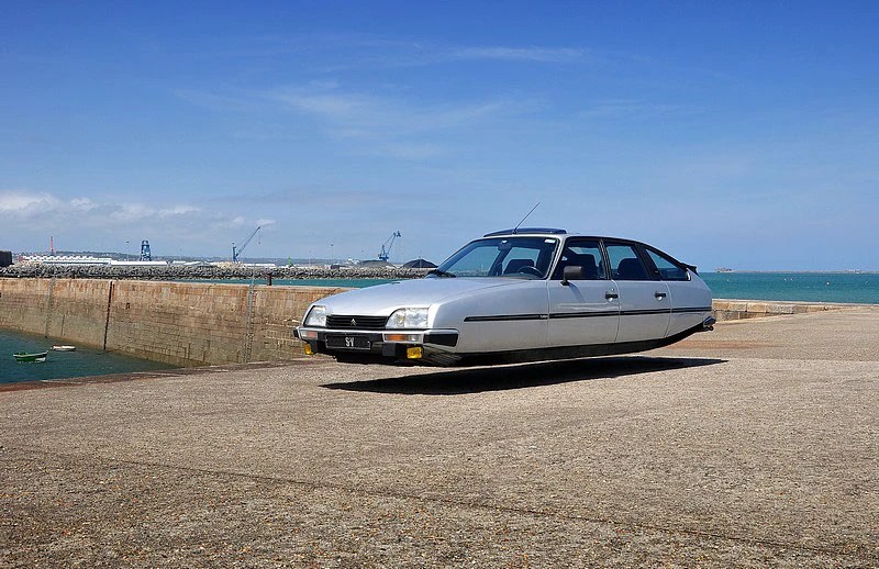 A silver floating car above a concrete surface near a harbor with a blue sky in the background.