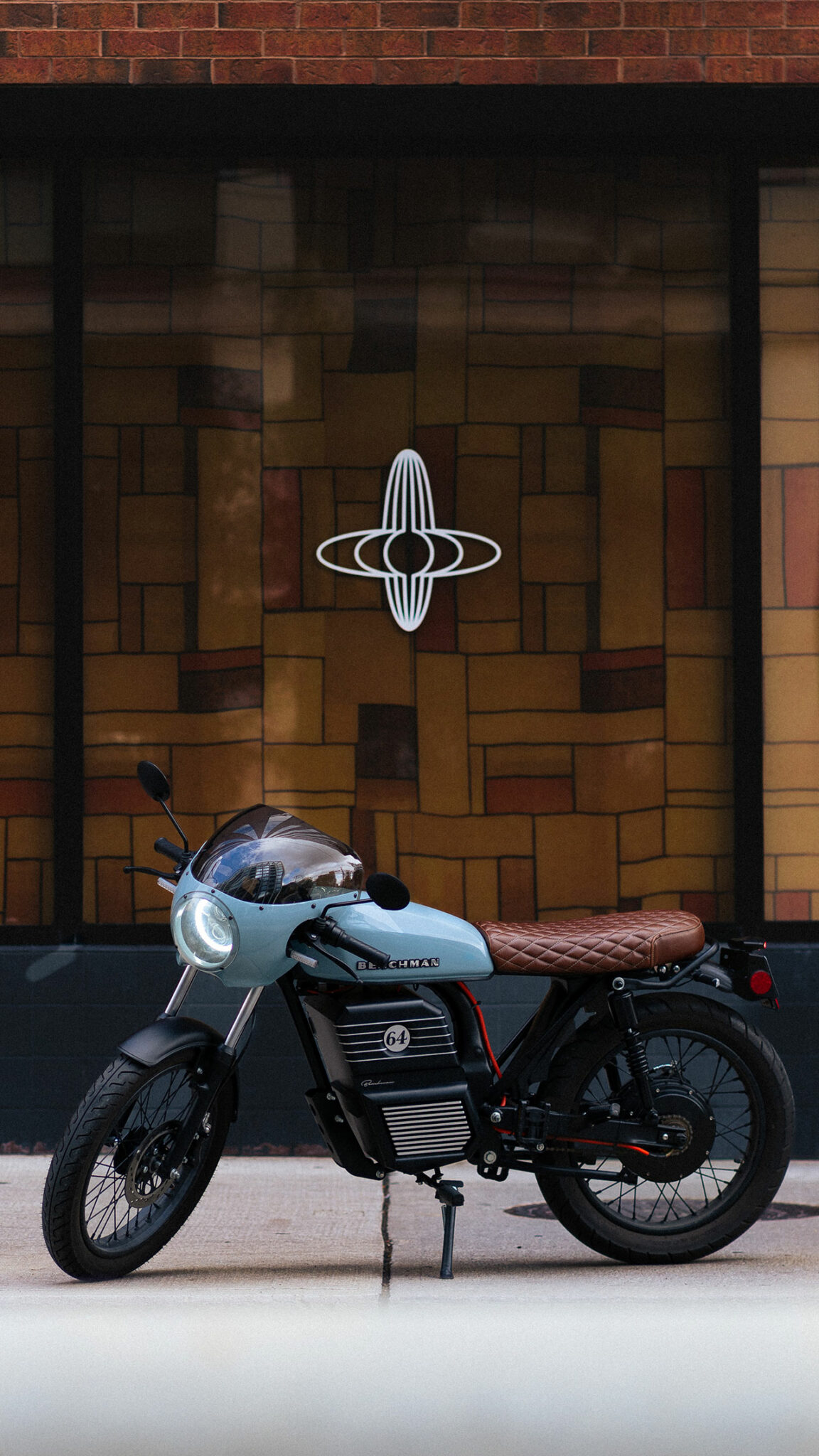 A retro-style electric motorcycle parked on the street, featuring a light blue body and a brown leather seat, with a distinctive logo visible on a brick wall behind it.