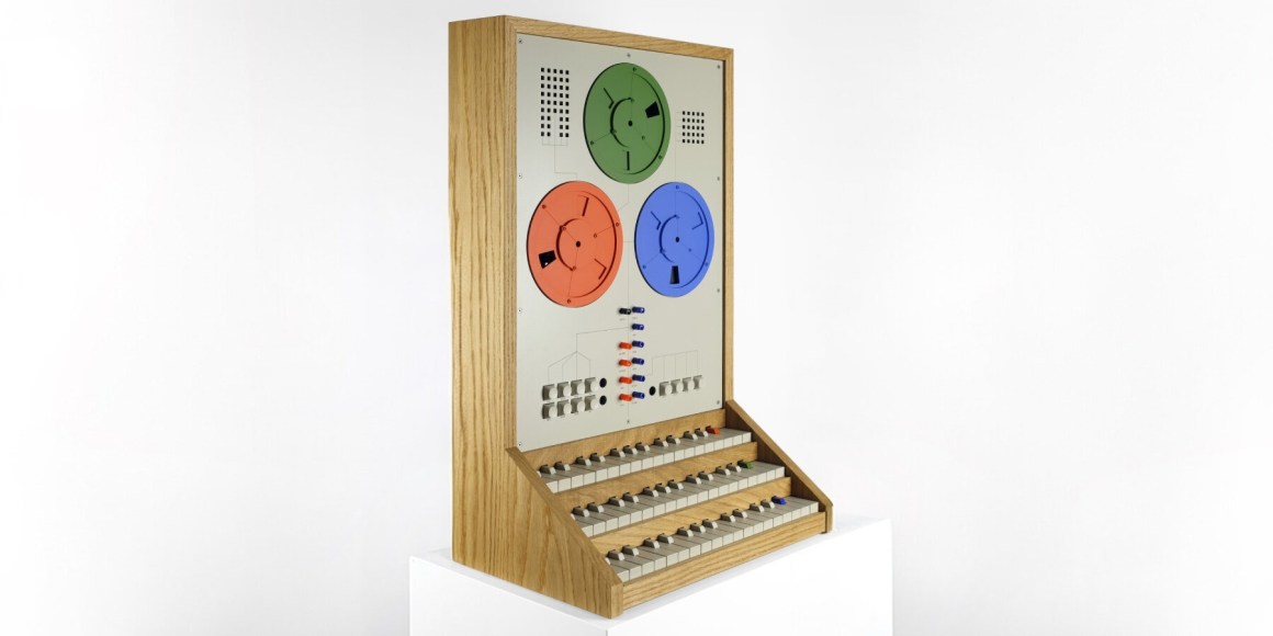 A vintage computer control panel featuring three large colored dials (red, green, and blue) and multiple knobs and buttons, set against a wooden frame.