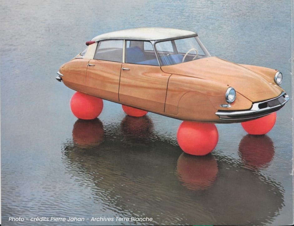 A vintage concept car with a unique design, featuring a wooden body and three large red buoy-like balls as wheels, floating on water.