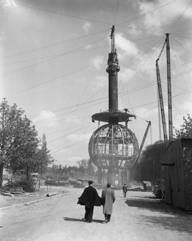 Vintage black and white photo of two men walking towards the construction site of the Atomium in Brussels, featuring a large spherical structure and cranes in the background.