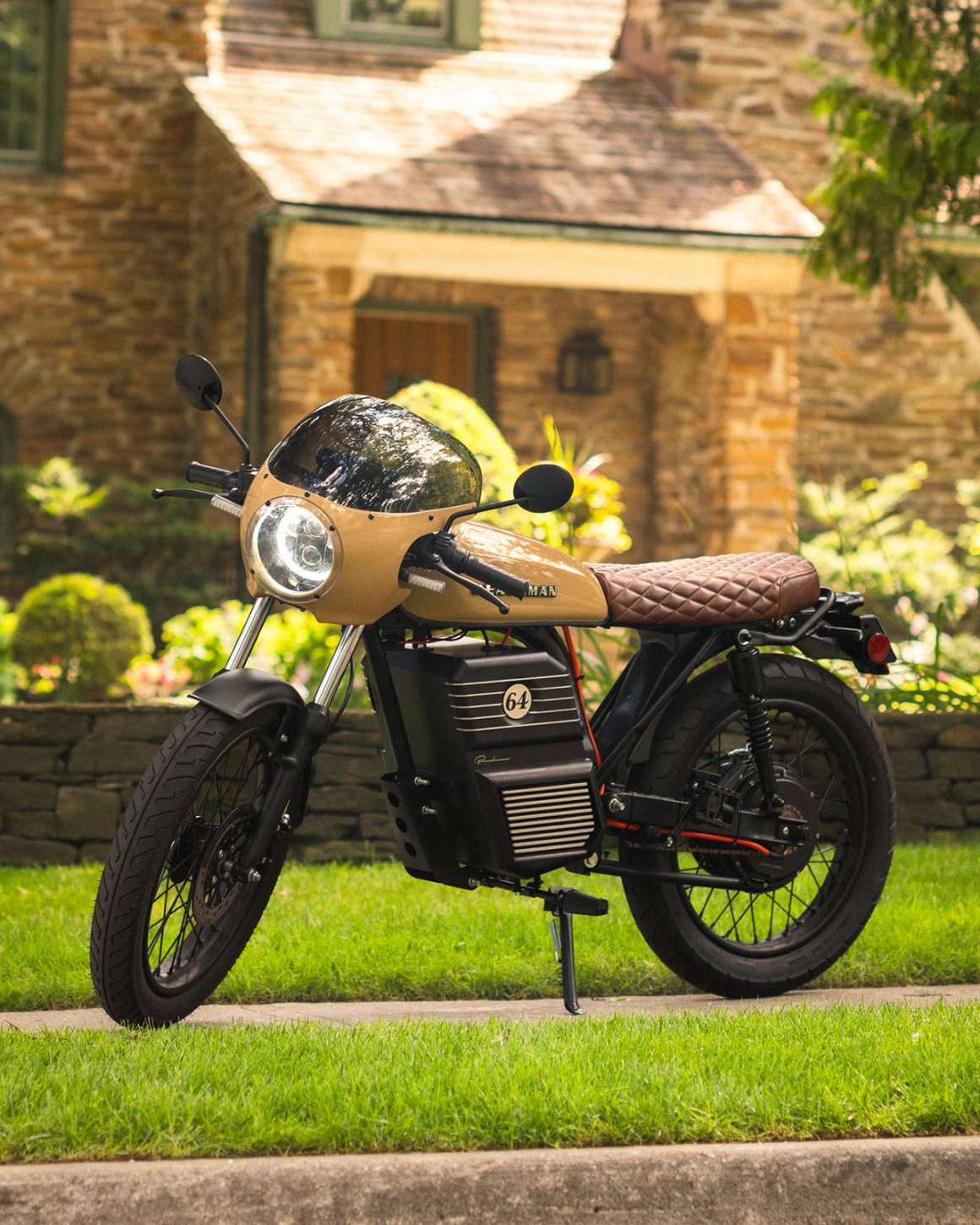 A stylish electric motorcycle parked on a grassy area in front of a stone house, featuring a tan body, a retro design, and a brown quilted seat.