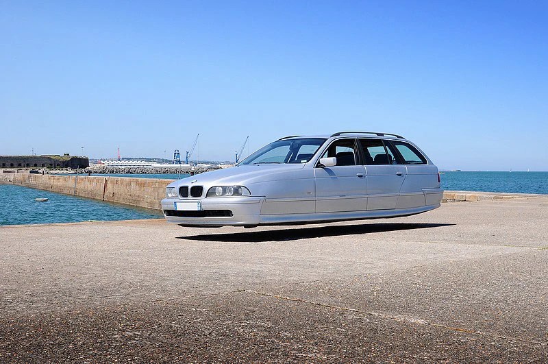 A digitally altered image of a silver car hovering above a concrete surface near a body of water, under a clear blue sky.