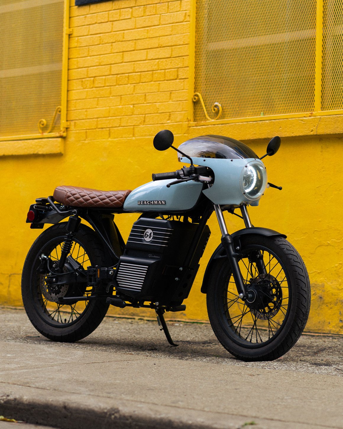 A stylish electric motorcycle with a light blue body and brown quilted seat parked against a vibrant yellow wall.