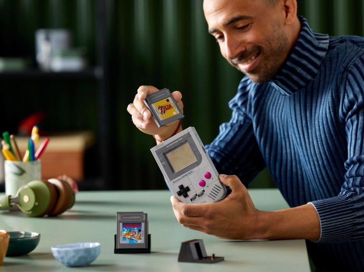 A person smiling while holding a retro handheld game console, preparing to insert a game cartridge into it, surrounded by colorful stationery and bowls on a tabletop.