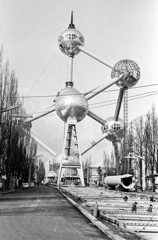 Black and white photograph of the Atomium under construction in Brussels, showcasing its unique spherical design and connecting tubes amid a developing urban landscape.