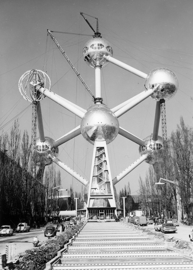 Black and white image of the Atomium in Brussels under construction, showing its unique structure with large spheres connected by tubes, cranes in the background, and a pathway leading to the entrance.