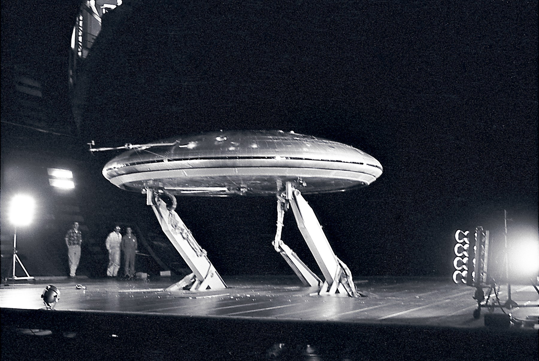 Black and white NASA photograph from March 1960 of the silver, disc-shaped Avrocar mounted on struts inside the enormous Ames wind tunnel, with three technicians standing beneath it