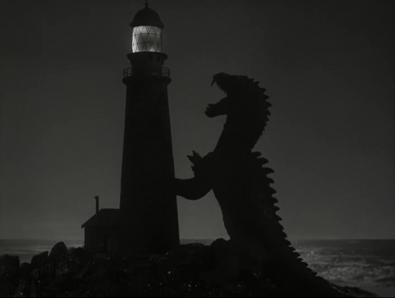 Black and white night scene from the trailer for The Beast from 20,000 Fathoms, the silhouetted spiny-backed Rhedosaurus rearing with jaws open against a tall lighthouse whose lantern room glows, dark sea and rocks below