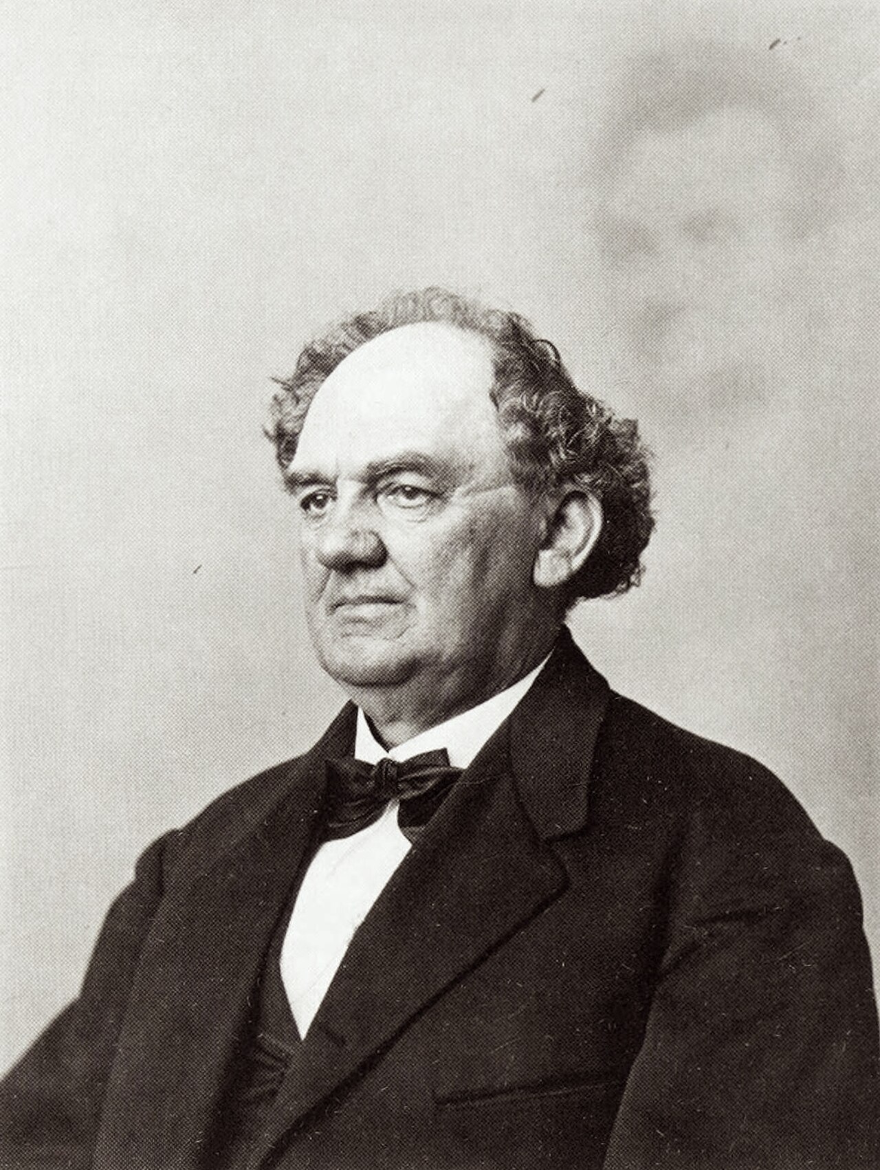 Black and white portrait photograph of P. T. Barnum, heavyset with curly gray hair, in a dark suit and bow tie, with a faint ghostly image of Abraham Lincoln's face hovering in the upper right of the background