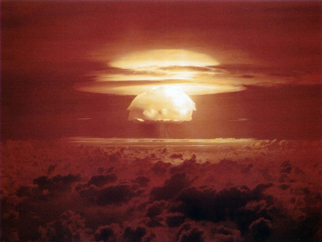 Color photograph of the Castle Bravo hydrogen bomb test, a glowing cream and gold mushroom cap ringed by flat condensation collars rising out of a blood red and rust orange sky above a dark cloud deck