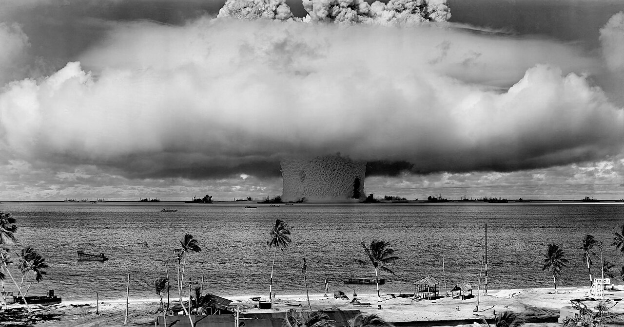Black and white panorama of the Crossroads Baker nuclear test, a vast white dome of condensation spreading over a dark column of water thrown up from the lagoon, with small target ships at its base and palm trees and huts on the beach in the foreground