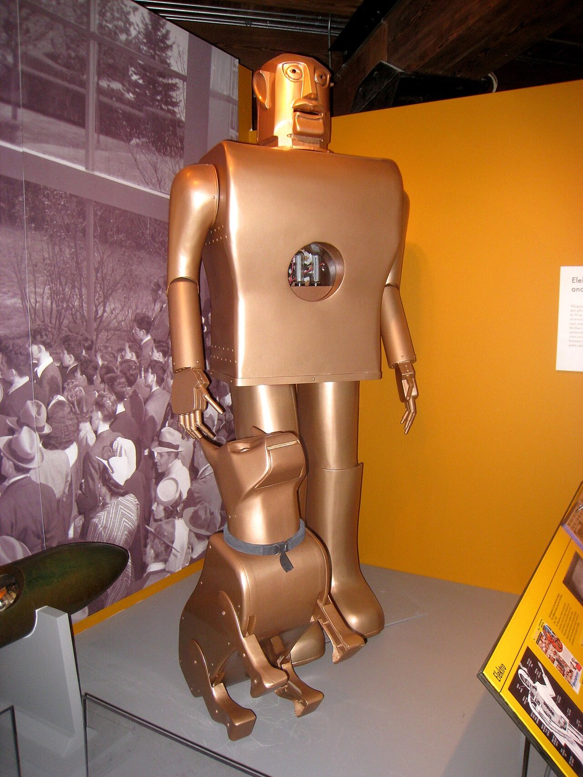Elektro, the seven-foot gold-colored Westinghouse robot from 1939, a blocky metal man with a round hole in its chest, standing in a museum display against an orange wall