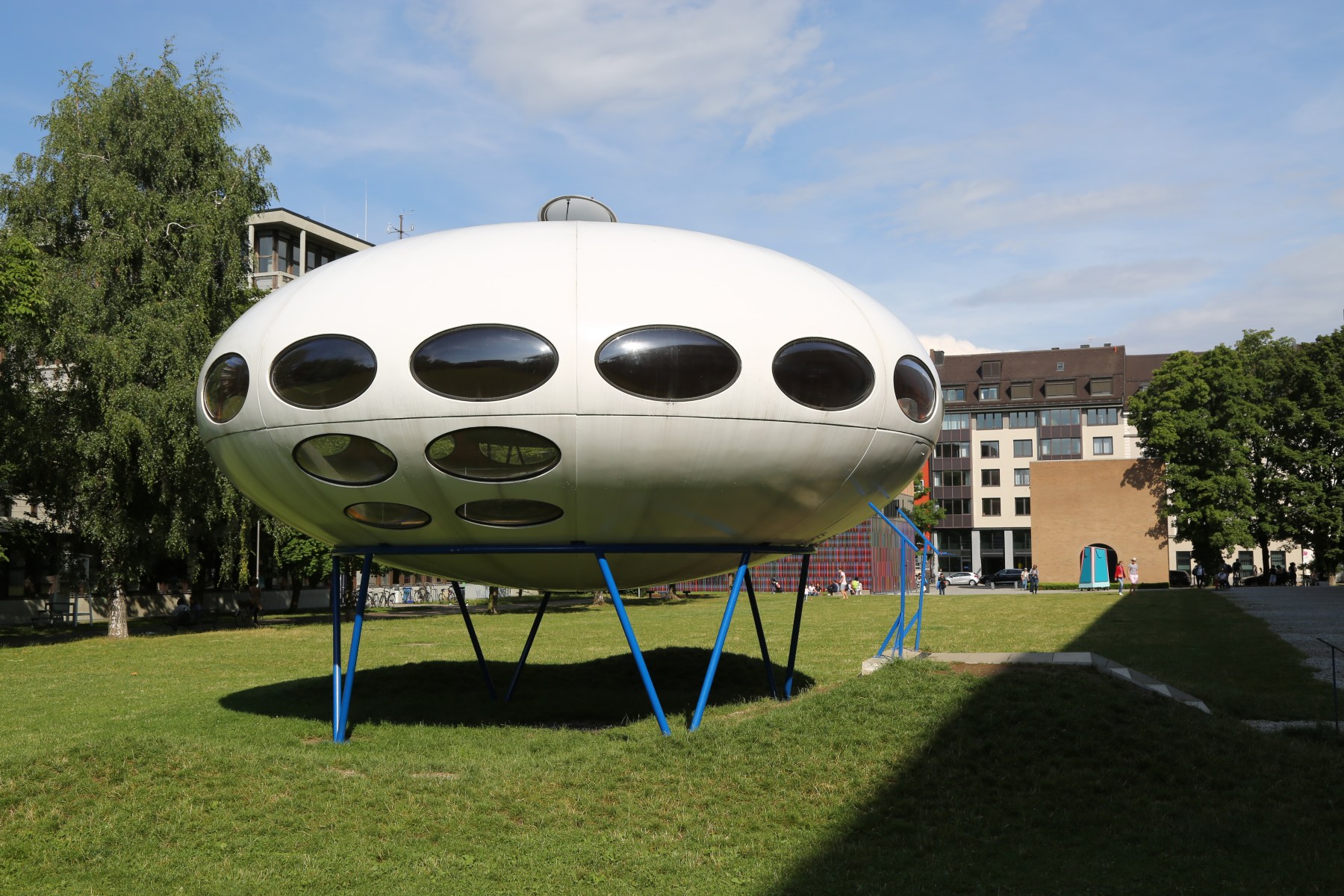 A white Futuro house, an ellipsoid plastic pod with a ring of oval windows standing on thin blue steel legs, on a lawn outside the Pinakothek der Moderne in Munich