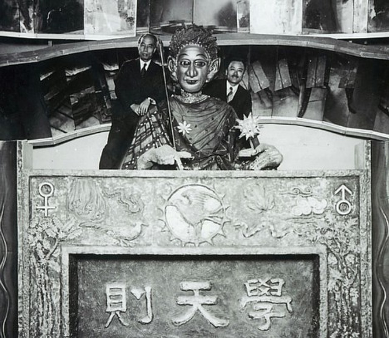 Black and white photograph from about 1928 of Gakutensoku, a large seated robot figure with a calm human face, a headdress and a robe, holding a pen and a lamp, set on an ornate carved desk with the characters 學天則, with two men in suits standing behind it