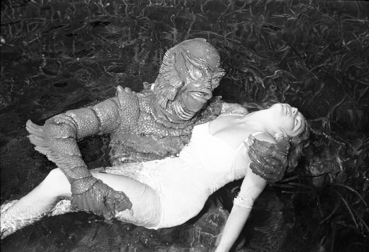 Black and white photograph from 1955 of the Gill-man lying in dark rippling spring water cradling the swimmer Ginger Stanley in a white swimsuit, her head thrown back, the scales and fin crest of the suit sharply detailed
