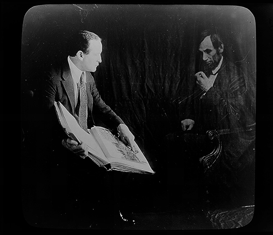 Black and white photograph of Harry Houdini seated in profile at left, holding open a large book, while a pale ghostly image of Abraham Lincoln appears to sit across from him at right against a dark background