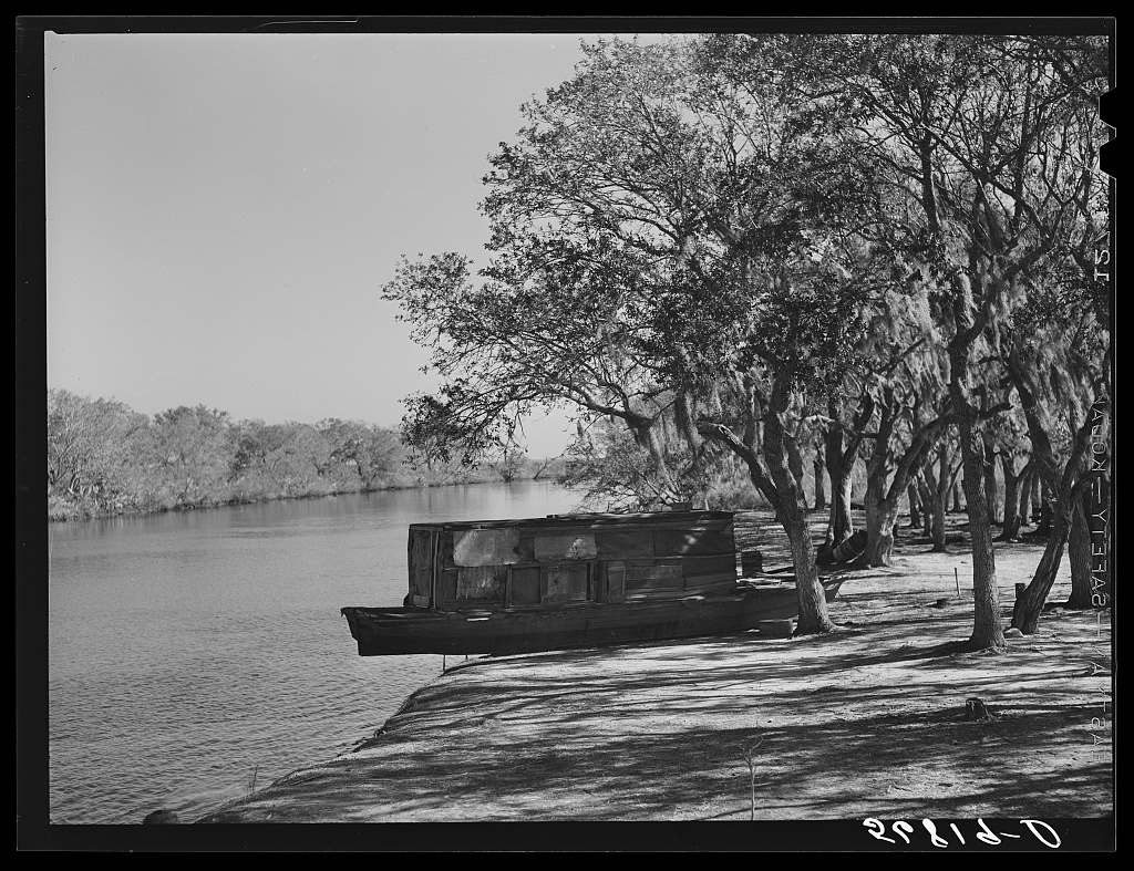 Black and white photograph by Marion Post Wolcott, 1941, of a wide Louisiana bayou on Delacroix Island with a beached houseboat and moss-hung oak trees casting dappled shade