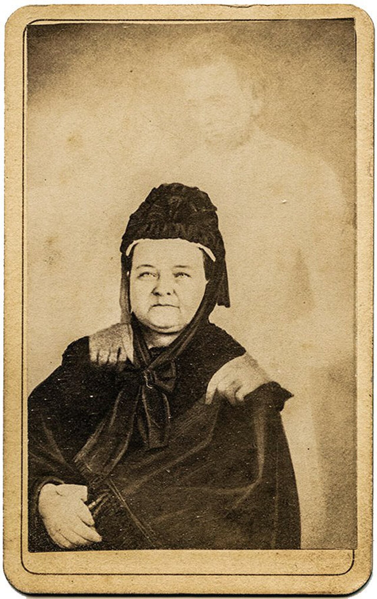 Sepia carte de visite of Mary Todd Lincoln seated in black mourning dress and bonnet, with the faint translucent figure of a tall bearded man resembling Abraham Lincoln standing behind her with his hands on her shoulders