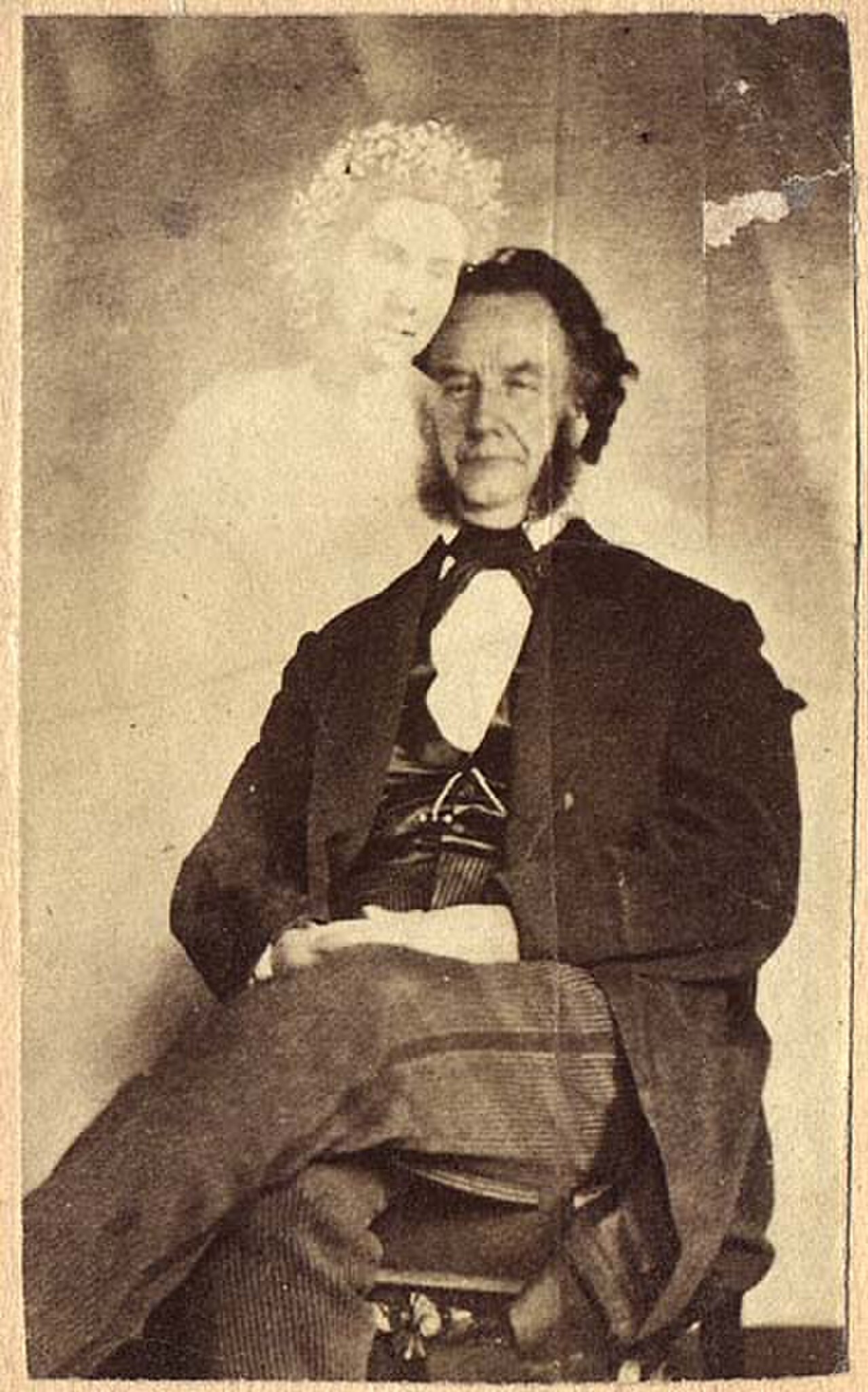Sepia carte de visite of Moses A. Dow, a seated bearded man in a dark suit, with the pale semi-transparent figure of a young woman wearing a wreath of flowers standing behind him and resting her hand on his chest