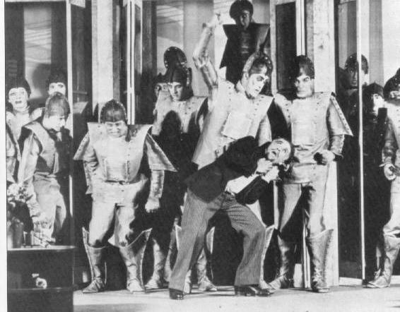 Black and white stage photograph from a 1928 production of R.U.R., a row of actors in padded robot costumes with tunics and boots looming over a man in a suit who staggers backward