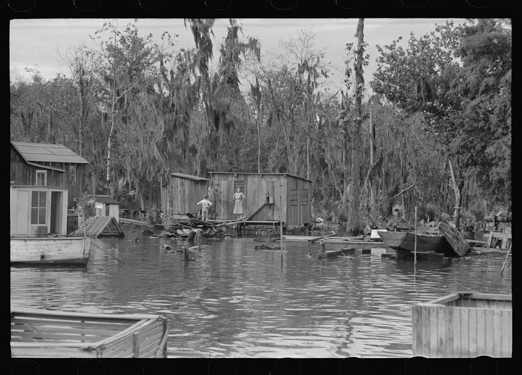 Black and white photograph by Russell Lee, 1938, of board shacks on stilts over still bayou water in Akers, Louisiana, with Spanish moss trees, skiffs and two figures on a porch