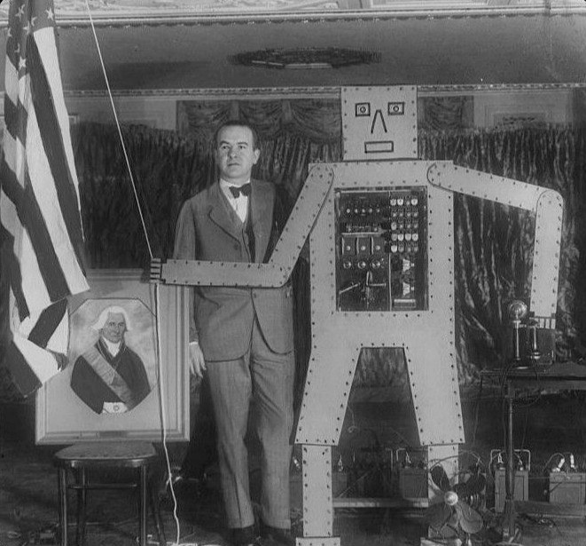 Black and white photograph from 1928 of R. J. Wensley in a suit standing beside Televox, a flat cut-out robot figure with a rectangular head, simple drawn face and a panel of switches and dials in its chest, one stiff arm raised