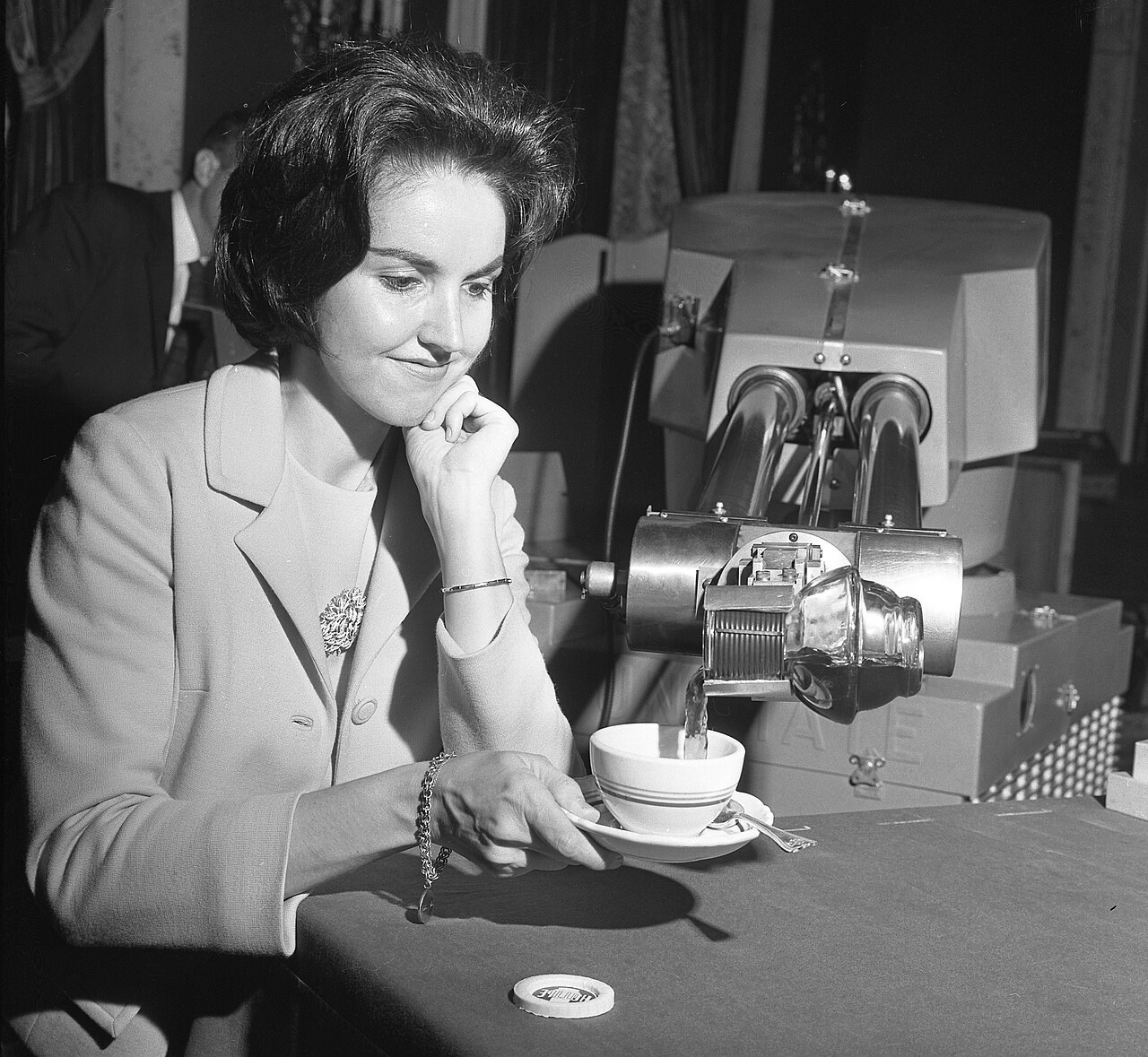 Black and white photograph from 1967 of a smiling woman seated at a table as the mechanical gripper arm of a large Unimate industrial robot pours coffee into her cup