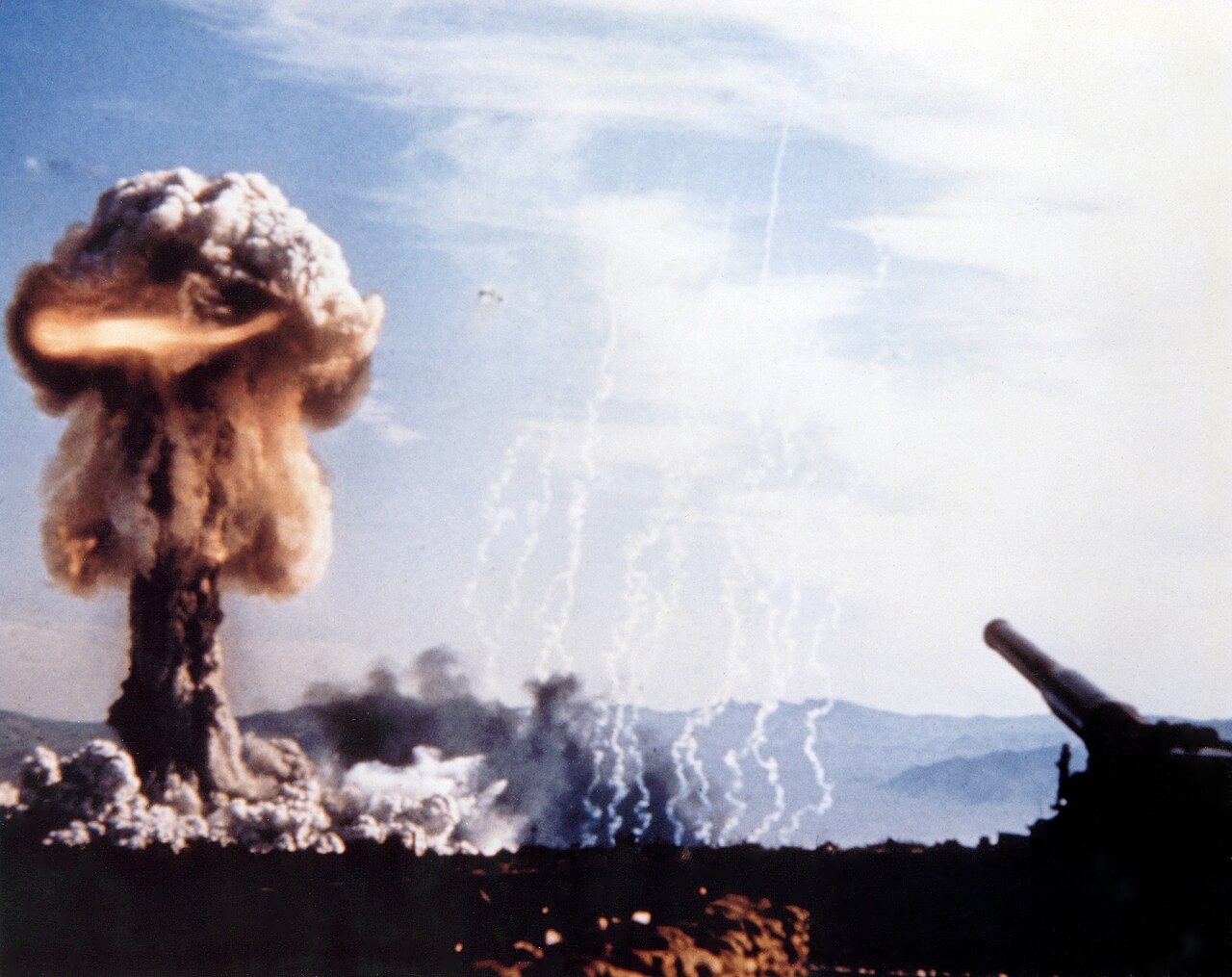 Color photograph of the Upshot-Knothole Grable test, a brown and orange mushroom cloud boiling up over the desert ringed by dust, with the long barrel of the 280mm atomic cannon jutting in from the lower right under a blue sky
